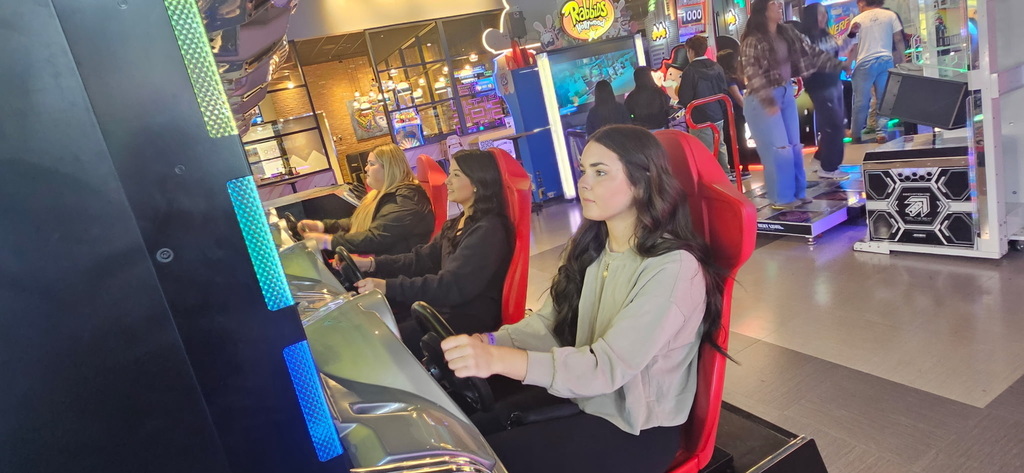 Three teenage girls sit in red seats, focusing intensely while playing car driving simulation arcade games.