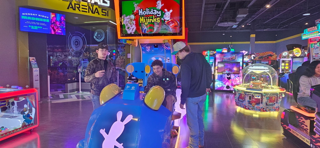 Three young men stand around a large, colorful children's racing arcade game in an area with neon lights
