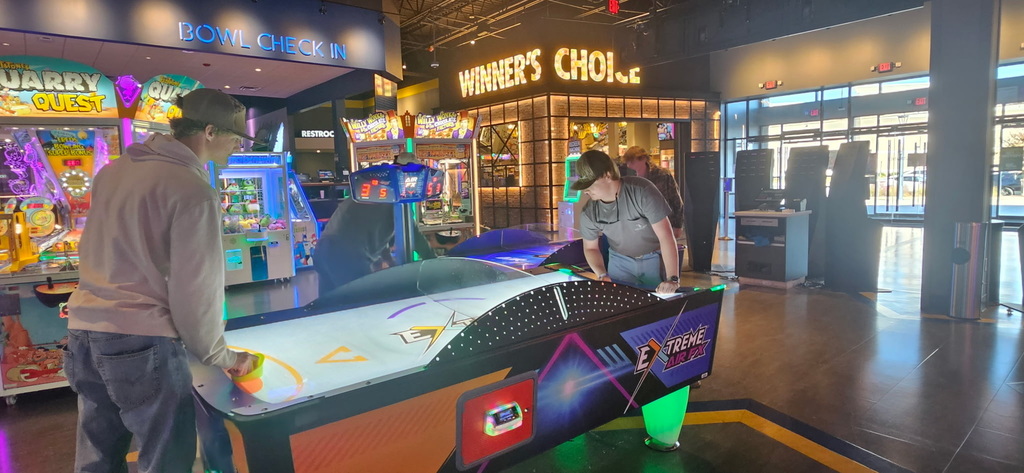 Two young men play an air hockey game in an arcade near the "Winner's Choice" prize counter and bowling check-in.