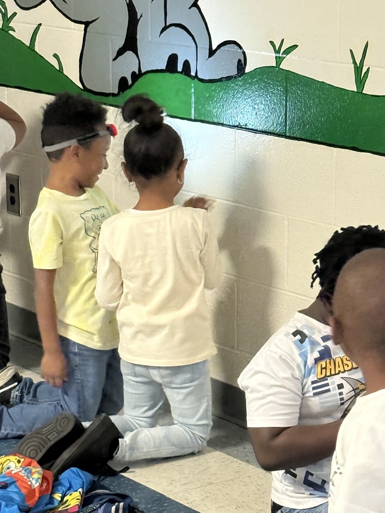 Children stand and kneel along a hallway wall, using their hands to make shadow shapes such as birds and animals. A bright flashlight creates crisp shadows on the white wall beneath a painted green mural.
