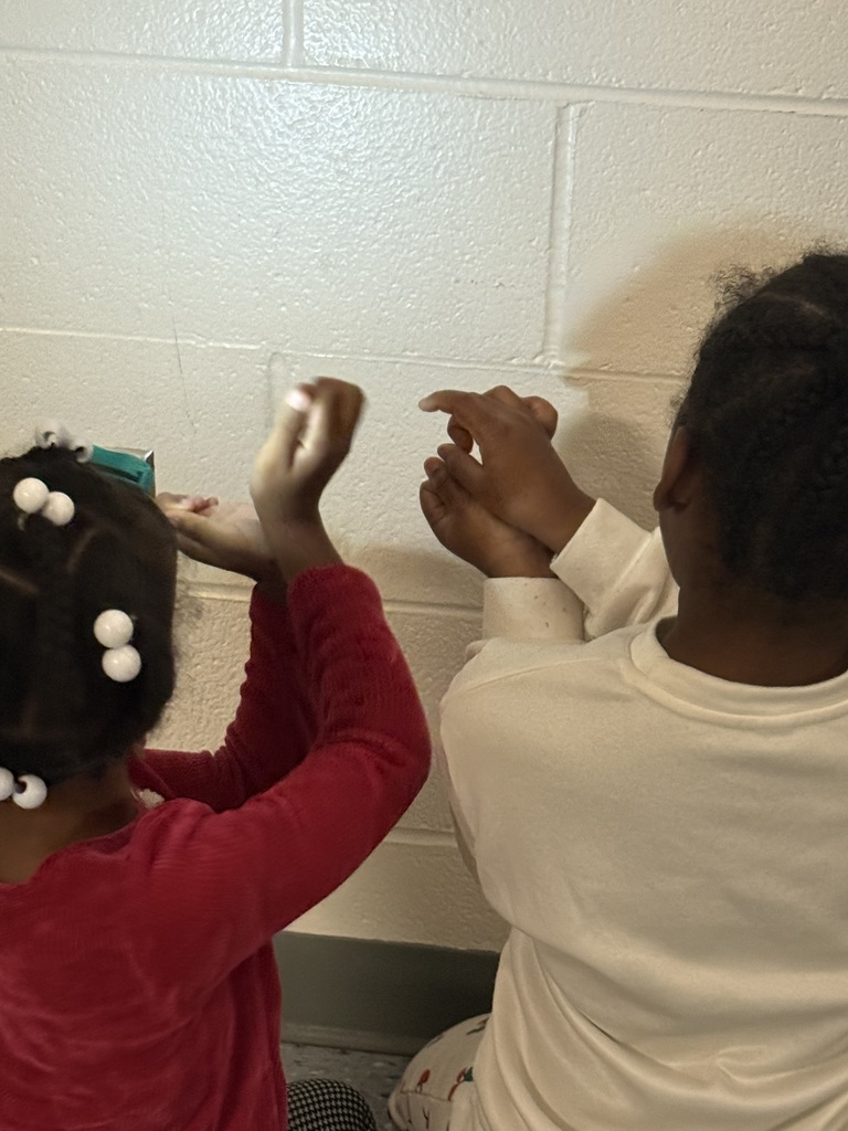 Two children sit in front of a wooden bookshelf, holding paper shadow puppets on sticks—shapes like sharks and sea creatures. A flashlight shines on the white cinderblock wall, creating clear shadows of their puppets. The students lean forward, experimenting with making the shadow grow and shrink.