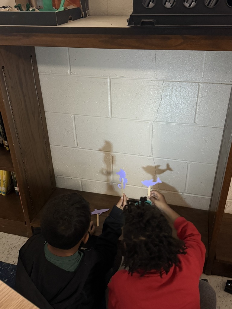 Two children sit in front of a wooden bookshelf, holding paper shadow puppets on sticks—shapes like sharks and sea creatures. A flashlight shines on the white cinderblock wall, creating clear shadows of their puppets. The students lean forward, experimenting with making the shadow grow and shrink.