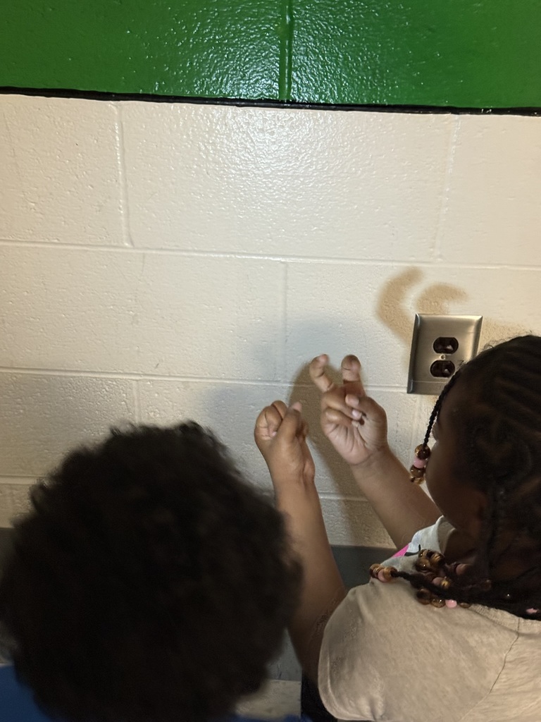 Two children kneel beside a white cinderblock wall, forming hand shapes in front of a flashlight to create animal-like shadow figures. Their shadows appear large on the wall as they experiment with bending fingers and changing hand positions.