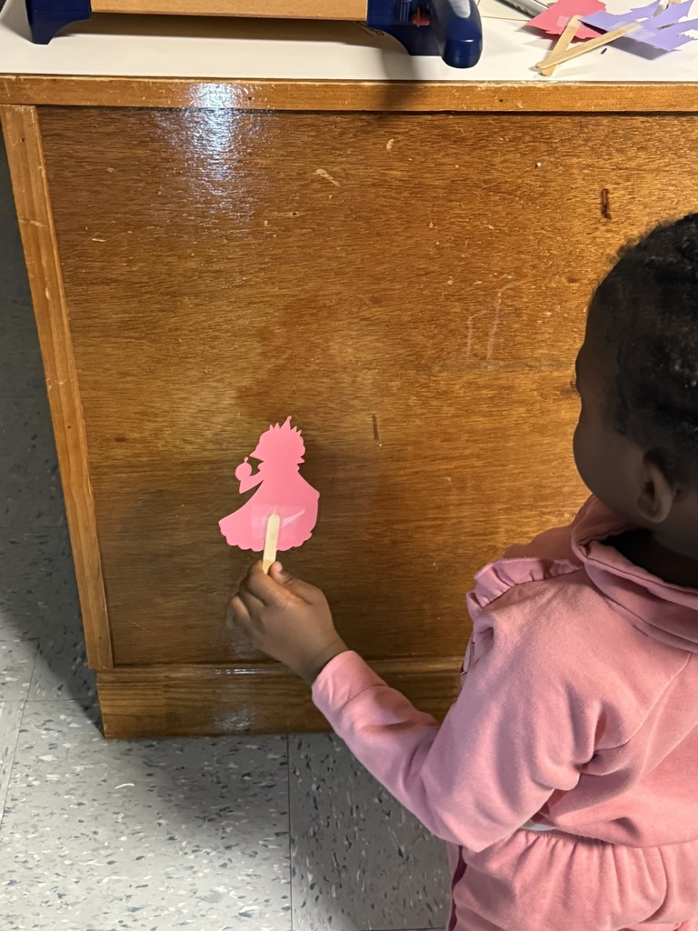 A young child in a pink outfit holds a pink paper princess cutout on a craft stick. The puppet’s shadow appears on the side of a wooden cabinet as the child explores how the flashlight changes the image.