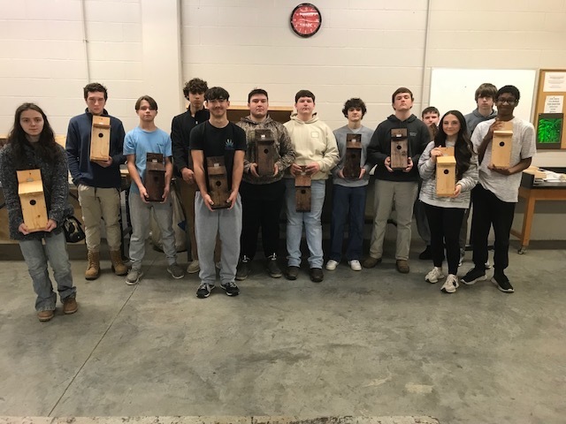 A group of high school students stand in a workshop, proudly holding the wooden birdhouses they built.