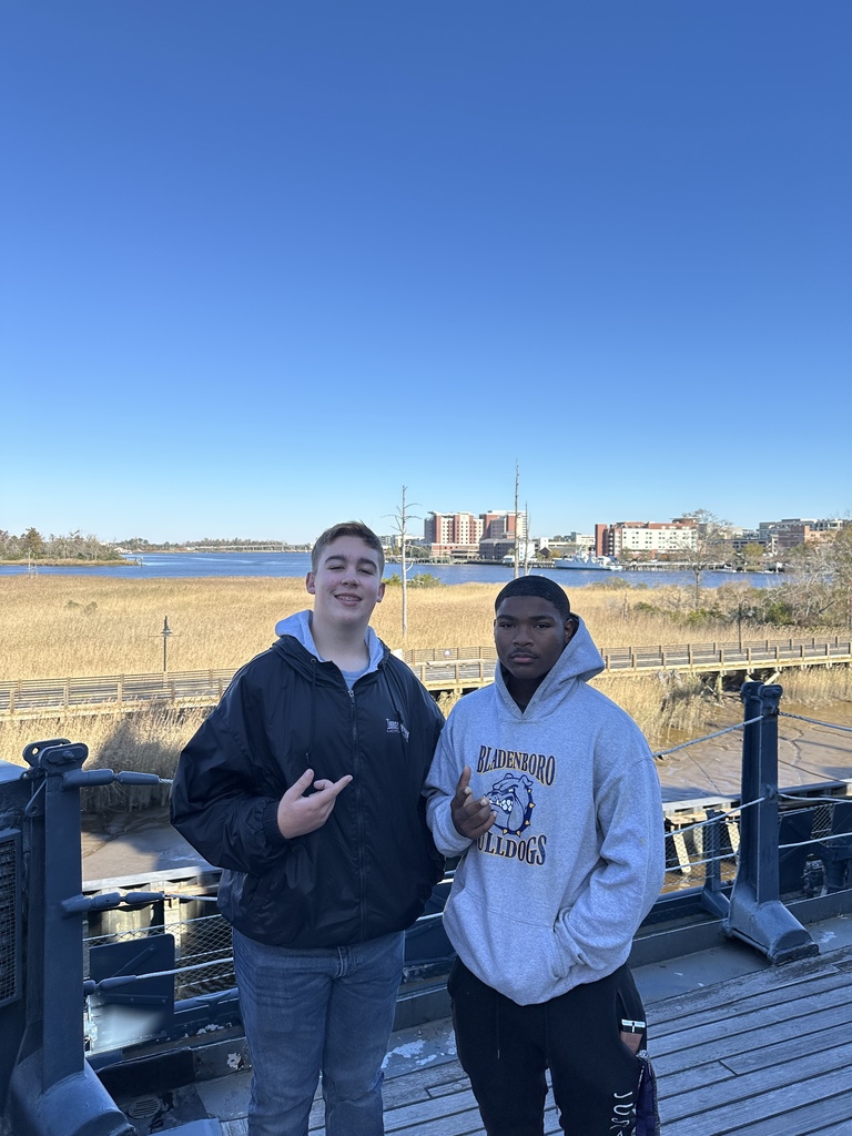 Two high school boys pose on the deck of a ship with a waterfront and city view in the background.