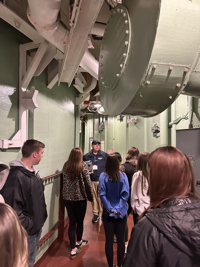 Students walk down a narrow hallway on a battleship while a guide gives a tour and explanation.