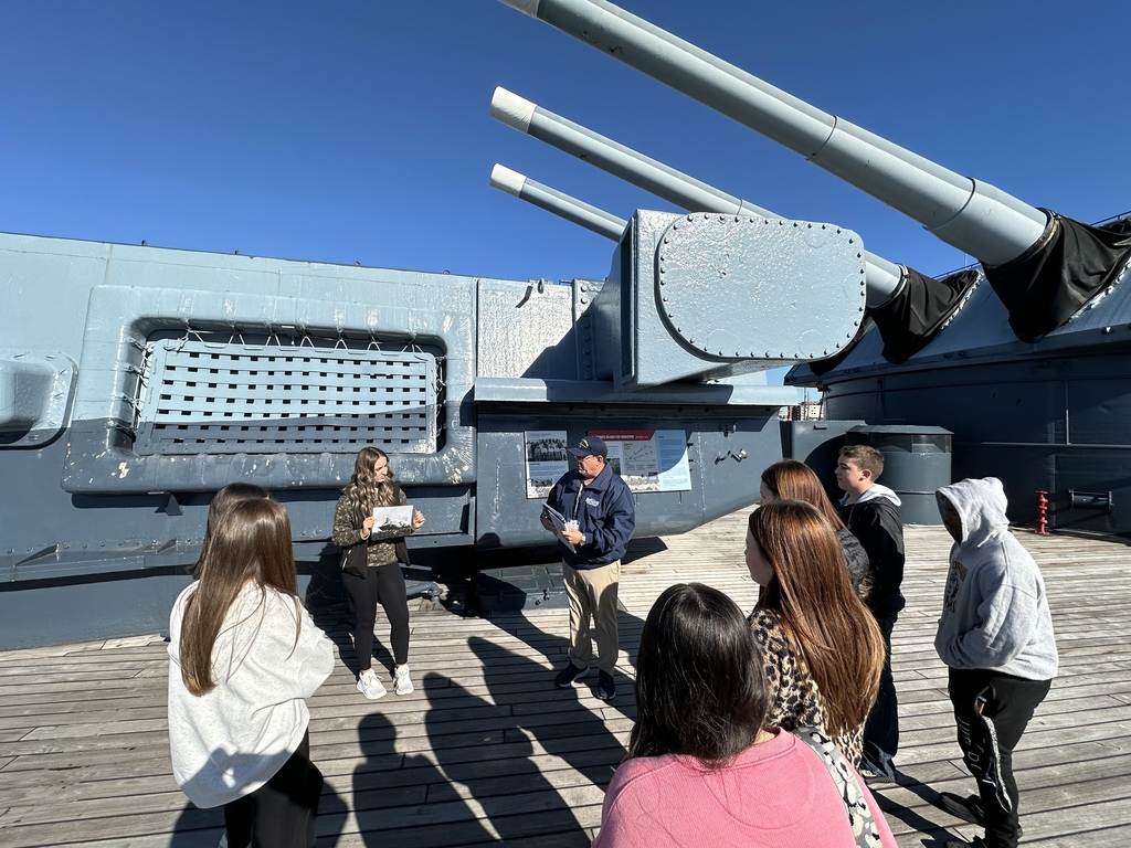 A group of students listen to a guide near the large main guns on the deck of a battleship.