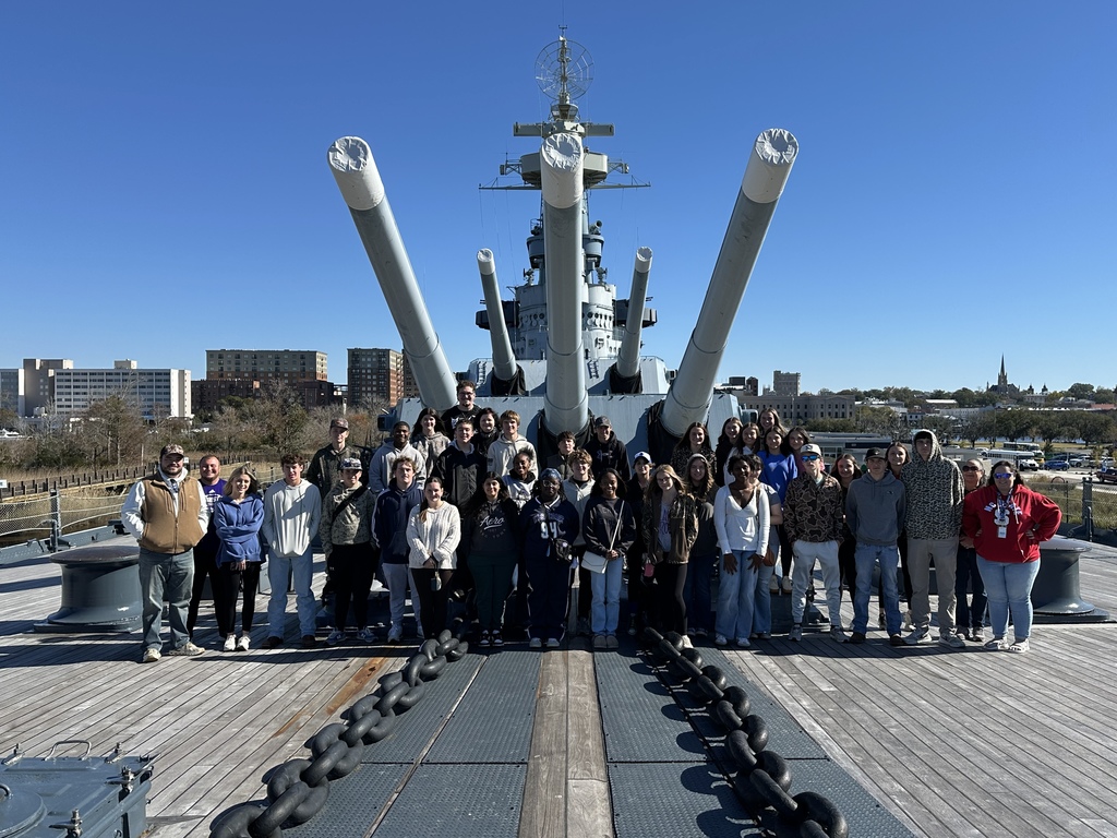 Large group of students and adults stand on a battleship deck with three large gun turrets behind them.