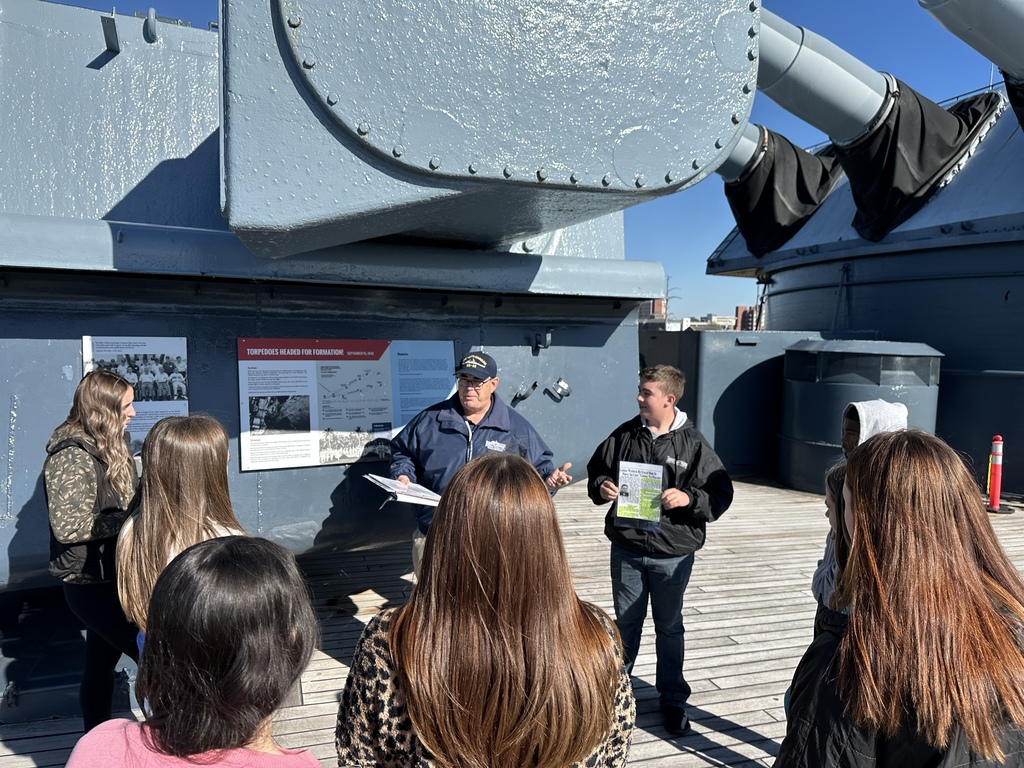 Students gather around a guide near a gun turret and informational sign on a battleship deck.