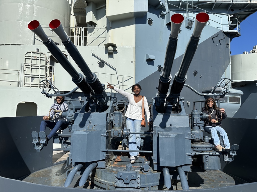 Three smiling students pose on or next to a dual 40mm anti-aircraft gun mount on a battleship