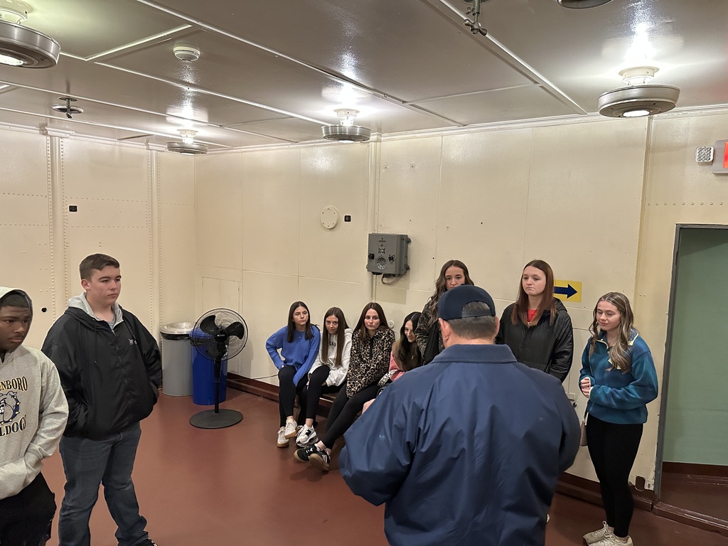 Students sit and listen to a guide giving a talk inside a white-walled room on a ship.