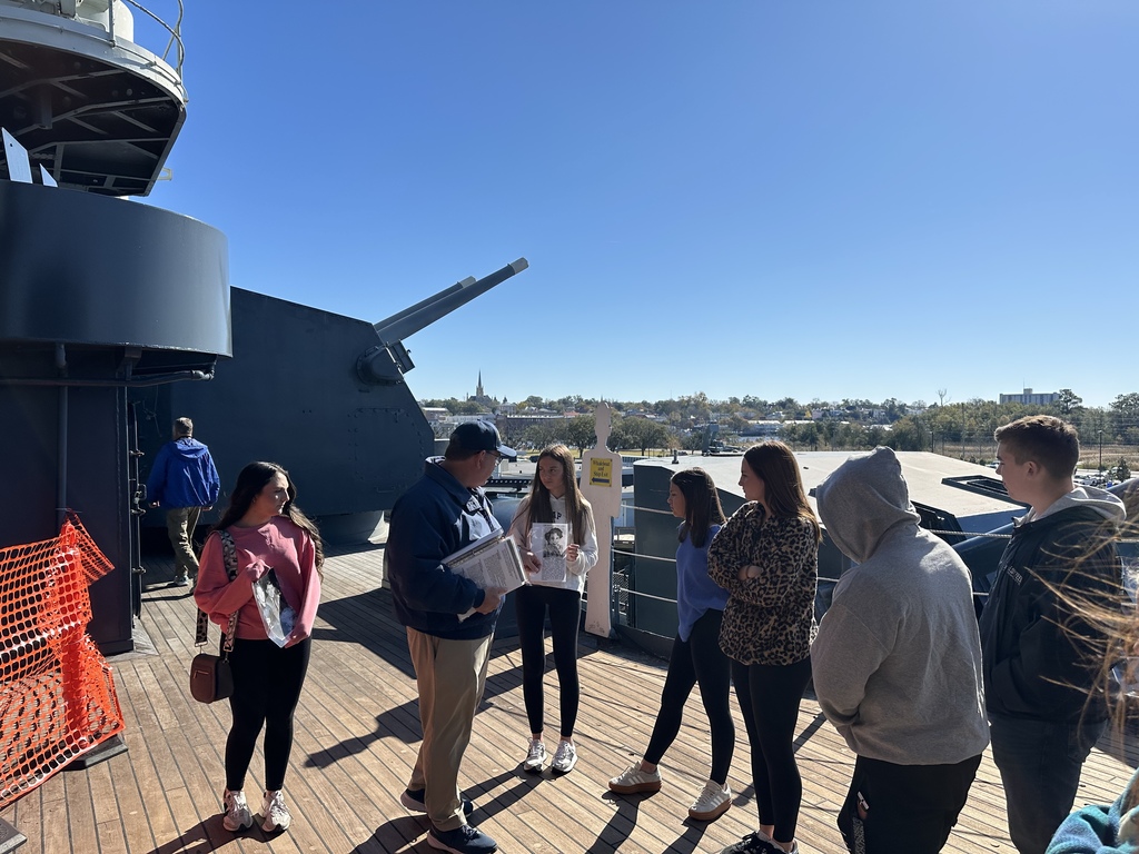 Students listen to a guide on the deck of a battleship with a gun turret and city view behind them