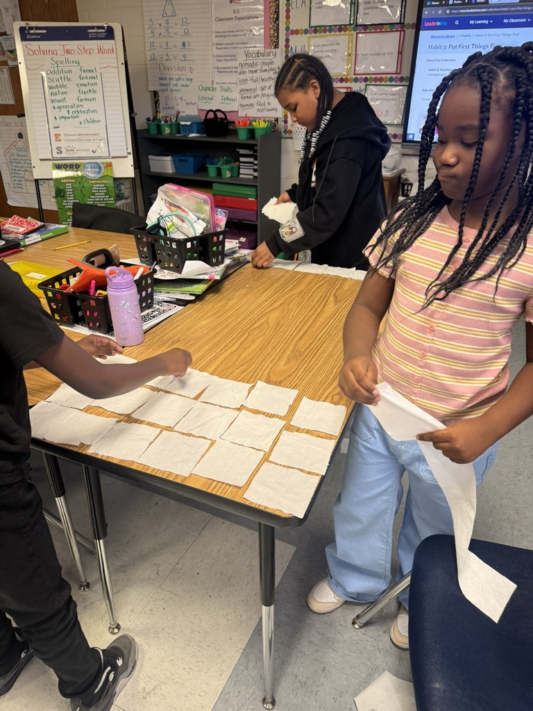 Three smiling students stand behind a long table fully covered in a grid of toilet paper squares. The boys pose proudly, and the classroom whiteboard and colorful posters are visible in the background.