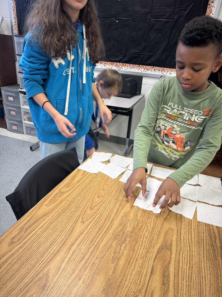 Two students stand at a desk arranging toilet paper squares into organized rows. One tears off more pieces while the other places them carefully on the tabletop.