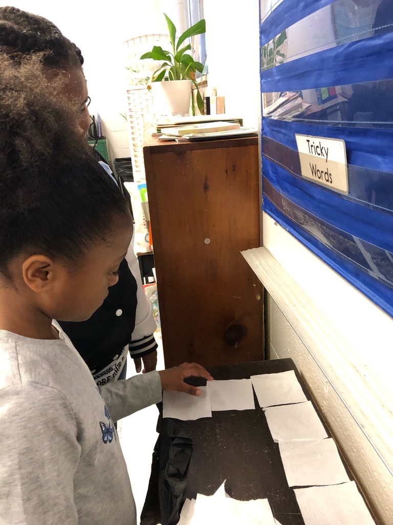 Two students place toilet paper squares on a small table near a pocket chart labeled “Tricky Words.” Another photo shows a student adding a square to a large table covered in evenly arranged toilet paper pieces.
