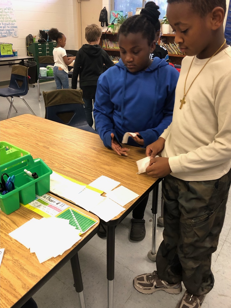 Two students stand at a classroom table arranging small squares of toilet paper in rows to measure area. Other students work in the background. Another photo shows two boys placing small squares onto a padded surface. A third photo shows three boys smiling behind a table fully covered with toilet paper squares arranged in a grid.