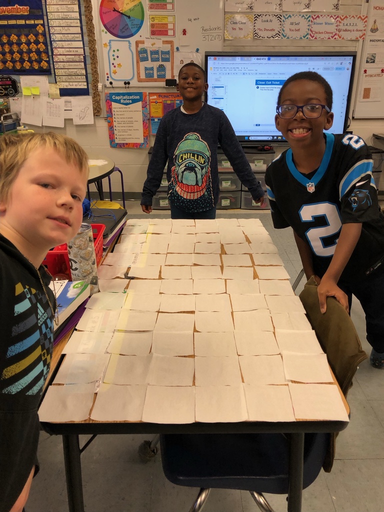 Three smiling students stand behind a long table fully covered in a grid of toilet paper squares. The boys pose proudly, and the classroom whiteboard and colorful posters are visible in the background.
