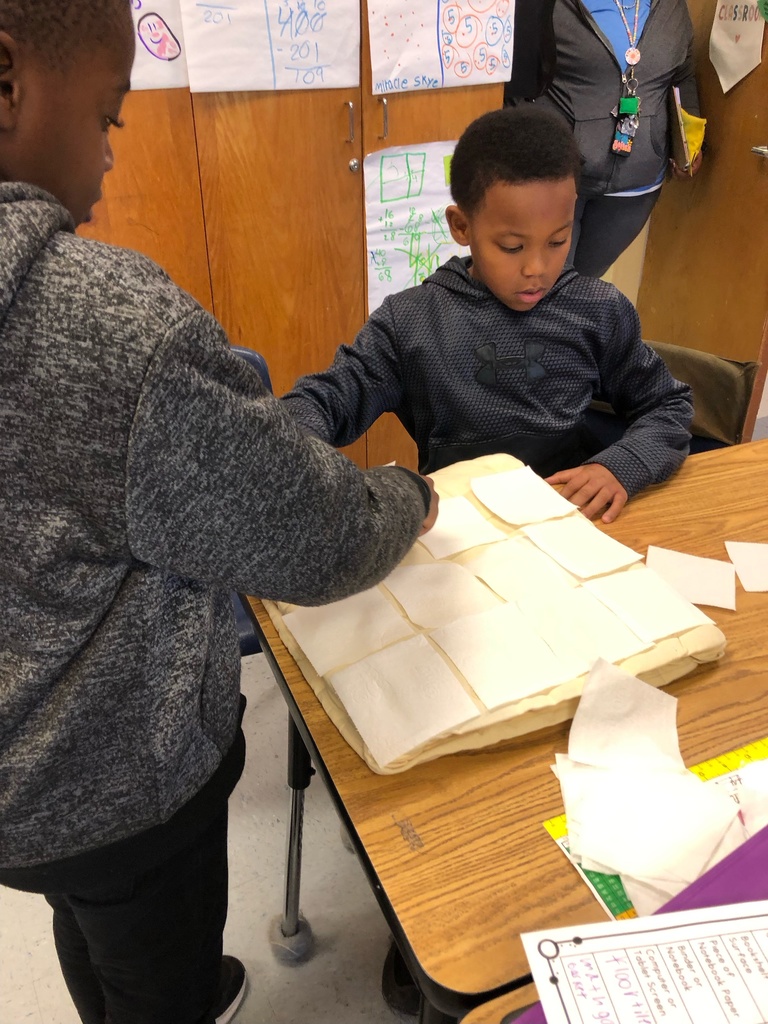 Two students place toilet paper squares on a small table near a pocket chart labeled “Tricky Words.” Another photo shows a student adding a square to a large table covered in evenly arranged toilet paper pieces.
