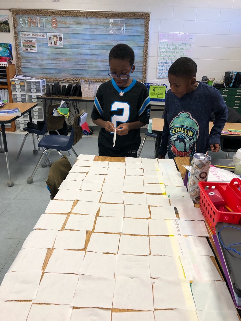 A student wearing a football jersey stands at a desk covered in toilet paper squares. Another student beside him watches as he prepares to add another piece to the grid.