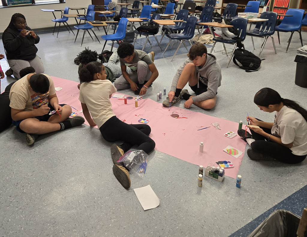 Students sit in small groups on the classroom floor painting a large pink poster, while others nearby cut out paper shapes and prepare materials.