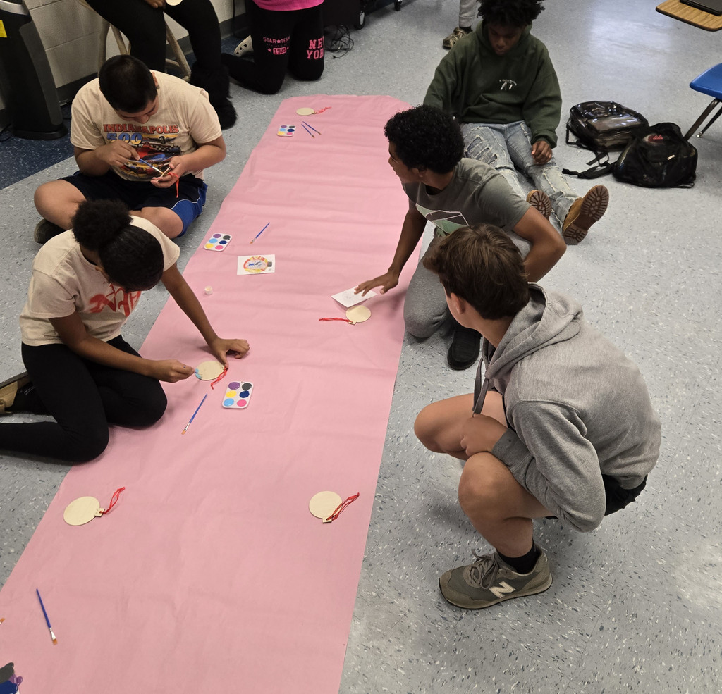 Students gather around a long sheet of pink paper on the classroom floor, painting small wooden ornaments while others stand nearby preparing supplies.