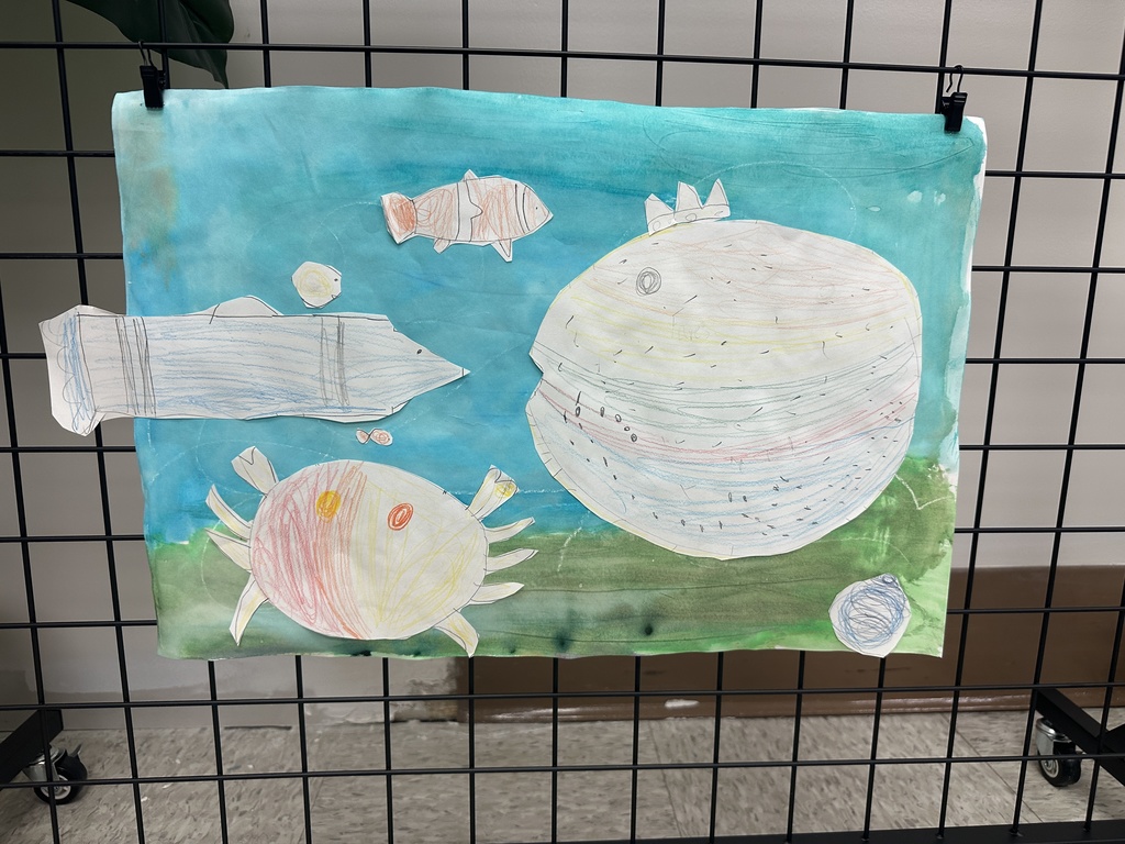 A student artwork of hand-drawn fish, a crab, and a large round sea creature placed on a blue watercolor ocean background.