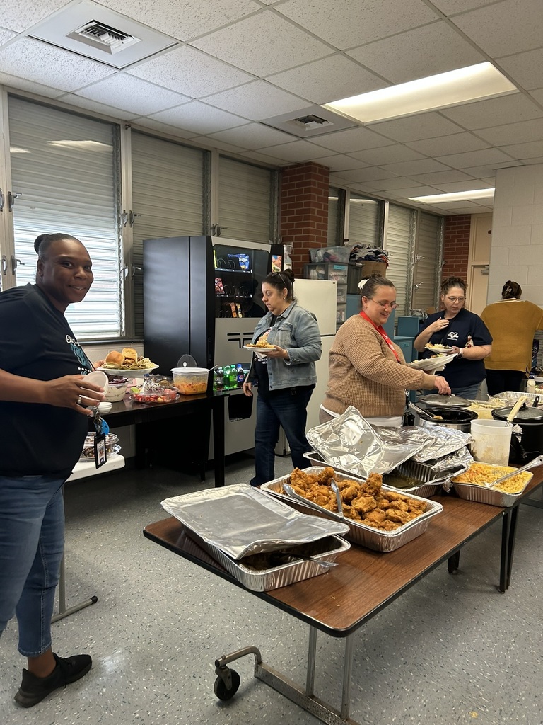 Staff members line up at a long table filled with aluminum trays of fried chicken, casseroles, and various dishes. One person smiles toward the camera while others scoop food. A vending machine and refrigerators are in the background, along with windows and storage shelves.