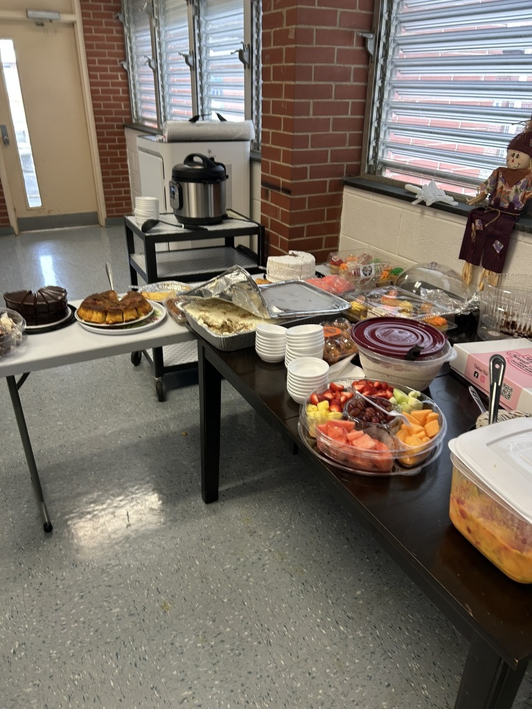 A table in the staff lounge is filled with desserts and fruit trays, including sliced melons, berries, cakes, and other dishes. There is an Instant Pot on a small cart and a scarecrow decoration sitting on the windowsill. The room has brick and white walls.
