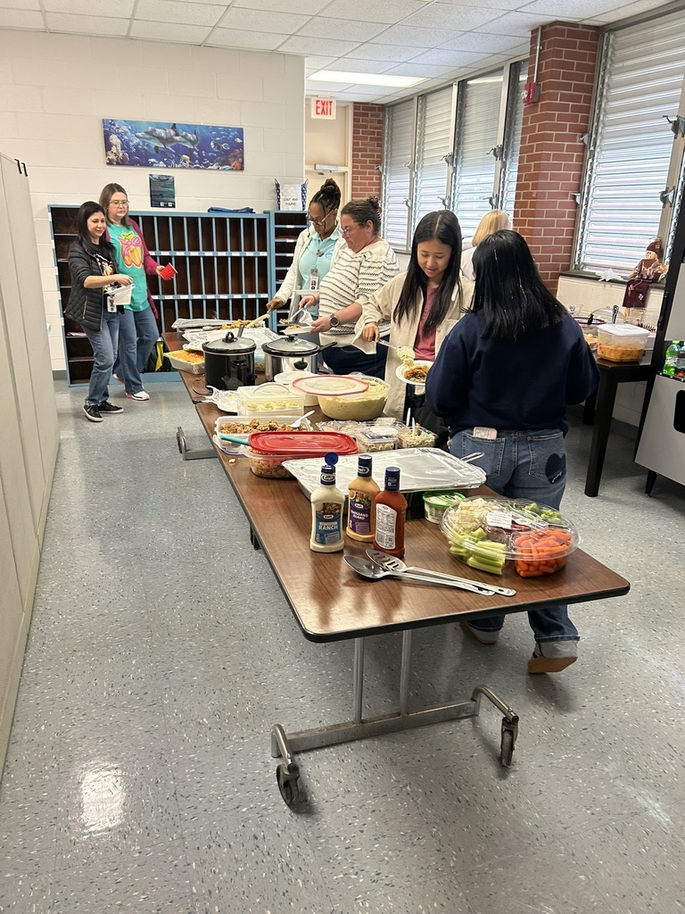 Several staff members stand in a serving line in the breakroom, filling plates with food from multiple crockpots, trays, and bowls on long tables. The room has brick walls, windows with metal blinds, and storage cubbies in the background. People are smiling and chatting as they serve themselves.