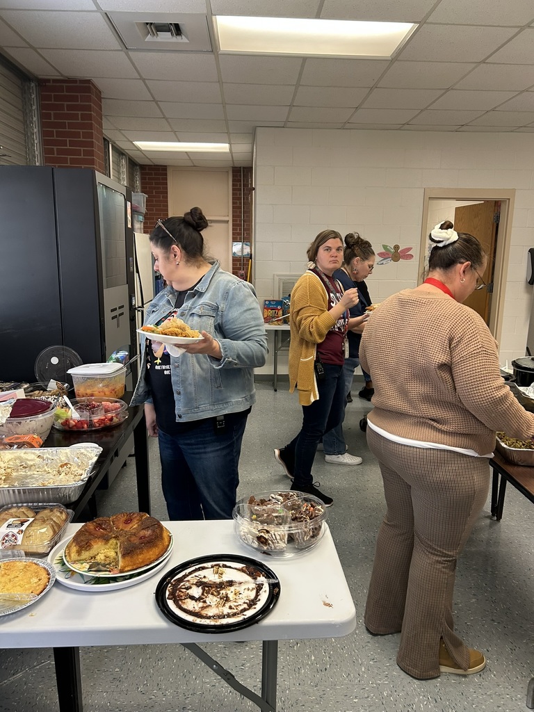 Staff members fill their plates from dessert trays and food dishes spread across several tables. The scene includes cakes, pies, casseroles, and trays of fruit. A vending machine, refrigerator, and open doorway are visible in the background.