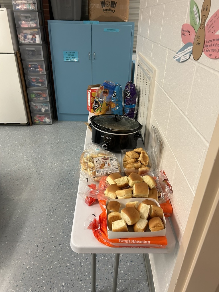 A small white table in a staff lounge holds a crockpot, bags of chips, Hawaiian rolls, and other bread items. Behind the table are blue storage cabinets, a stack of plastic drawer units, and a labeled cardboard box. The wall has student artwork displayed to the right.