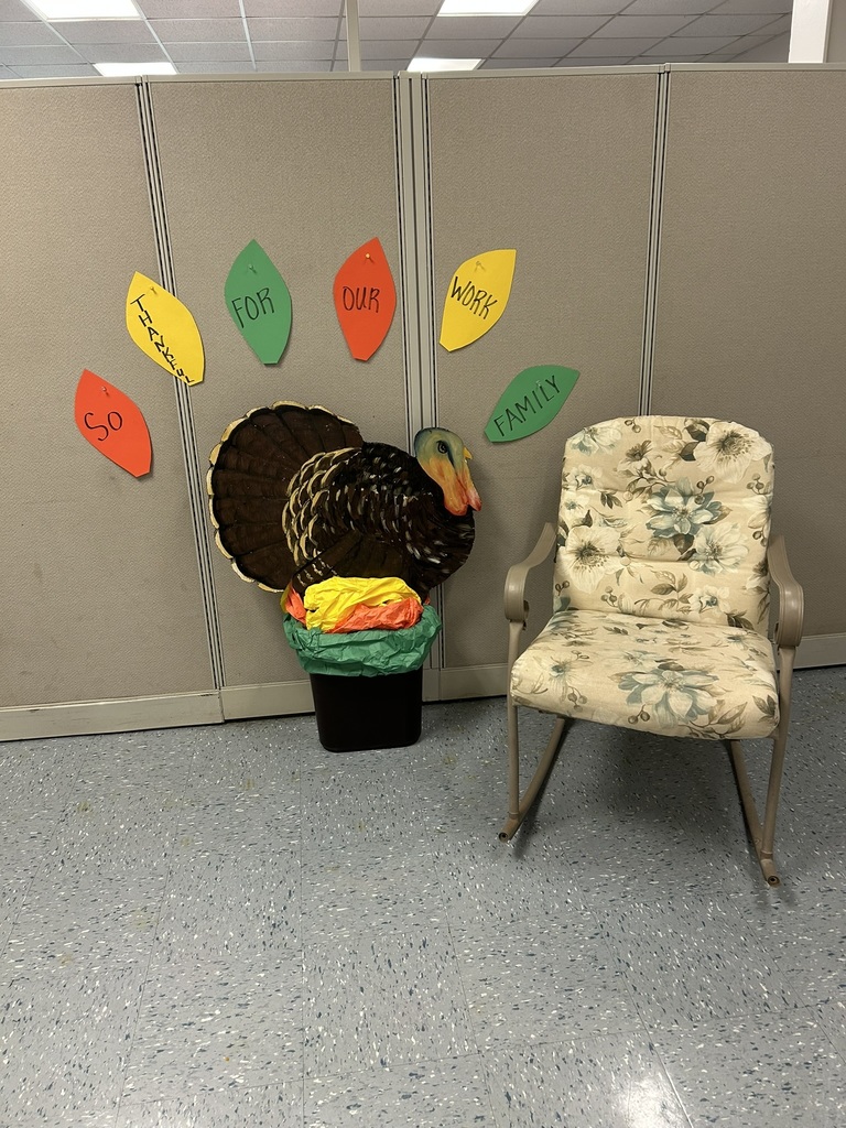A decorated office cubicle wall features a large paper turkey with colorful tissue paper at the base. Around the turkey, there are cut-out leaves in red, yellow, and green that read “So Thankful for Our Work Family.” A floral cushioned chair sits to the right of the display on a speckled blue linoleum floor.