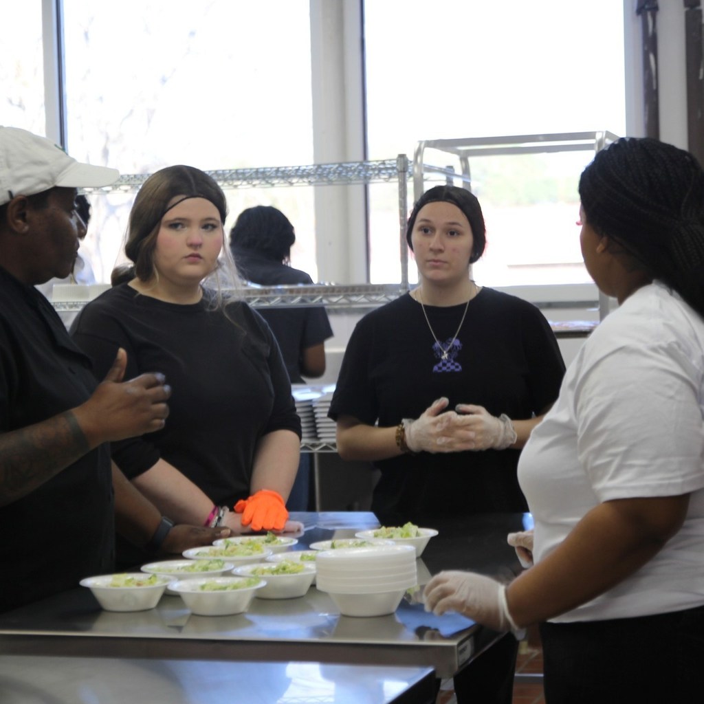 Students gather around a kitchen counter as an instructor explains food preparation steps.