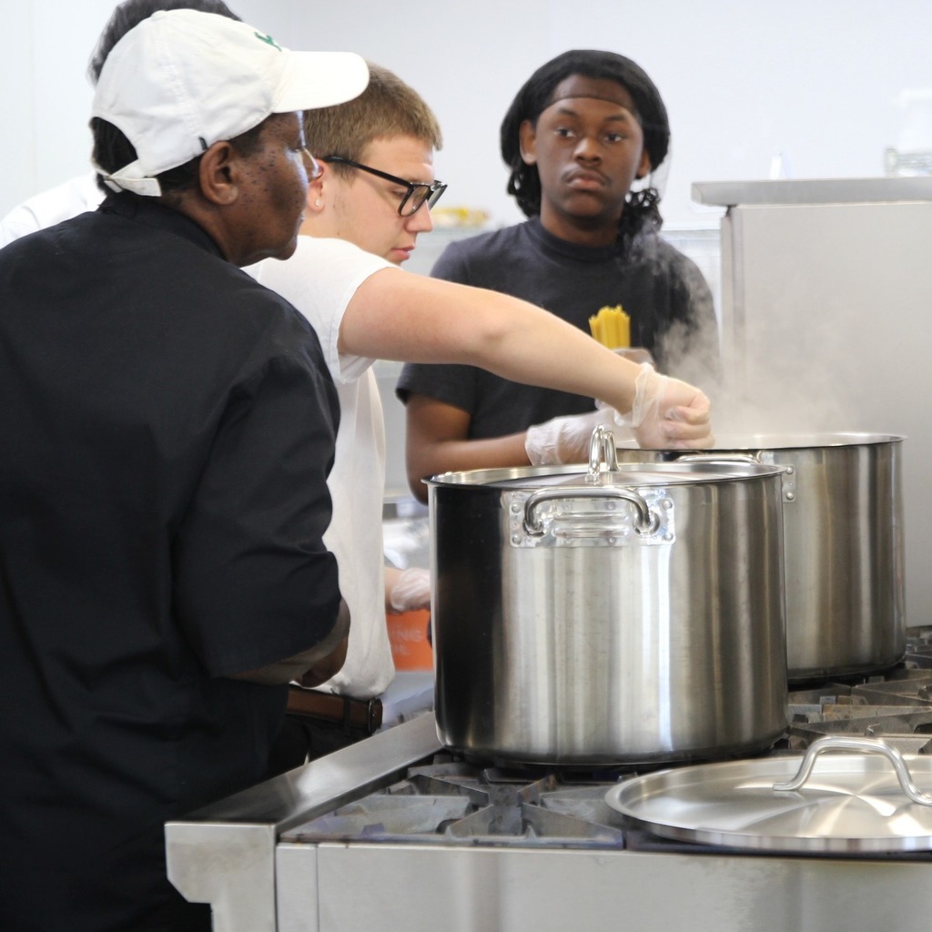 Students cook pasta with guidance from an instructor beside large steaming pots in a commercial kitchen.