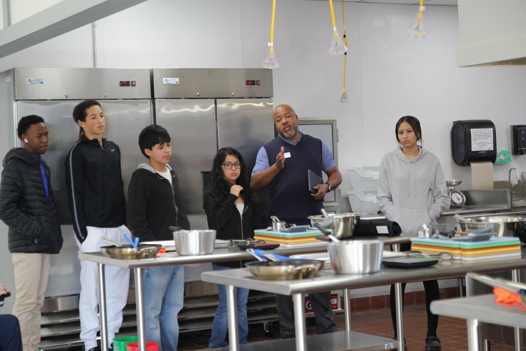 Students stand in a commercial kitchen listening to an instructor speak during a culinary demonstration.