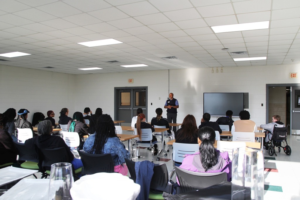 A speaker addresses a room of students seated at tables during an indoor presentation.