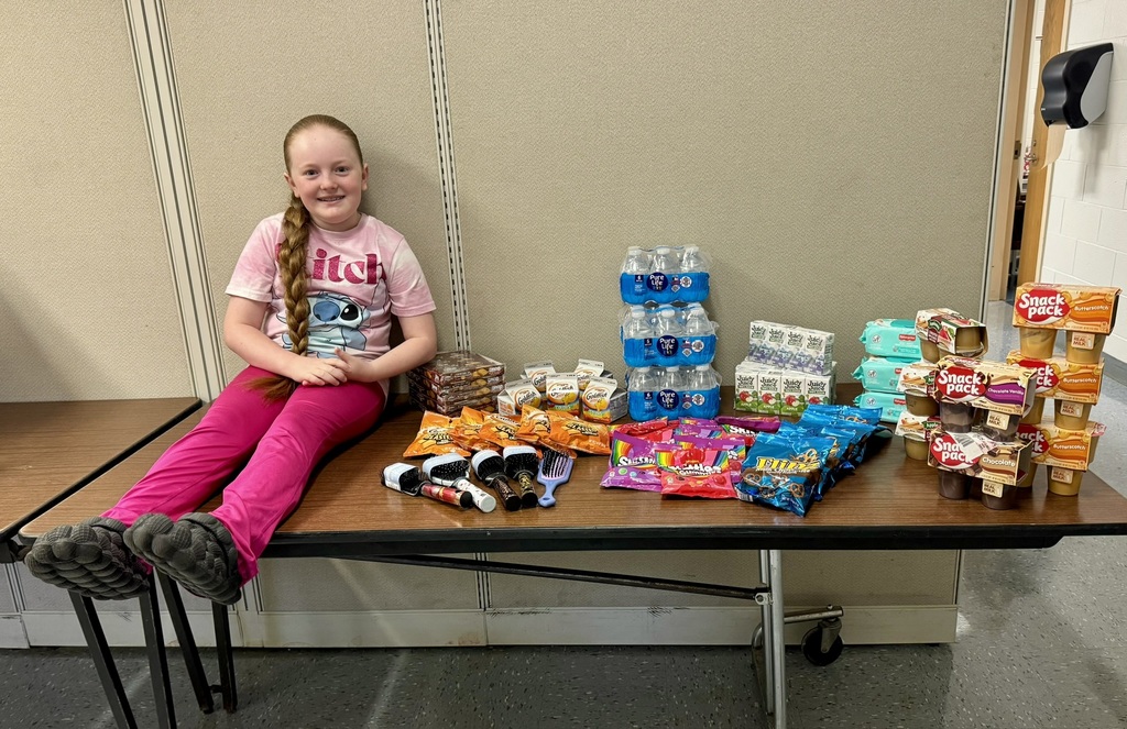 A young girl with long braided hair wearing a pink Stitch shirt sits on a table beside an assortment of donated items. The table is filled with water bottles, Juicy Juice boxes, Snack Pack puddings, Goldfish crackers, snack cakes, hairbrushes, socks, and candy. The background shows a beige folding partition wall.