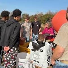 A diverse group of students and adults stands outdoors, gathered around bags of soil and equipment, engaged in a lesson
