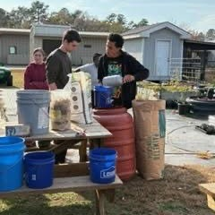 Two young men mix materials on a wooden table outdoors, surrounded by buckets, bags, and barrels.