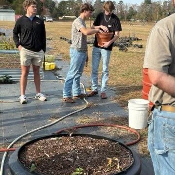 Three young men are standing in a garden area with one holding a plant pot.