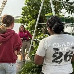 Two young women face an instructor in a greenhouse, surrounded by lush plants.