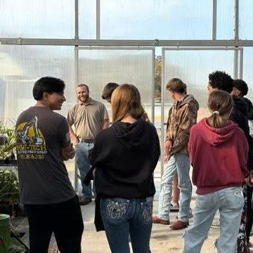 A group of students and adults gathers in a greenhouse, interacting and observing their surroundings.