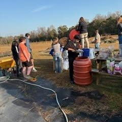 A diverse group of people gather outdoors, some tending to plants, with equipment and supplies scattered around.