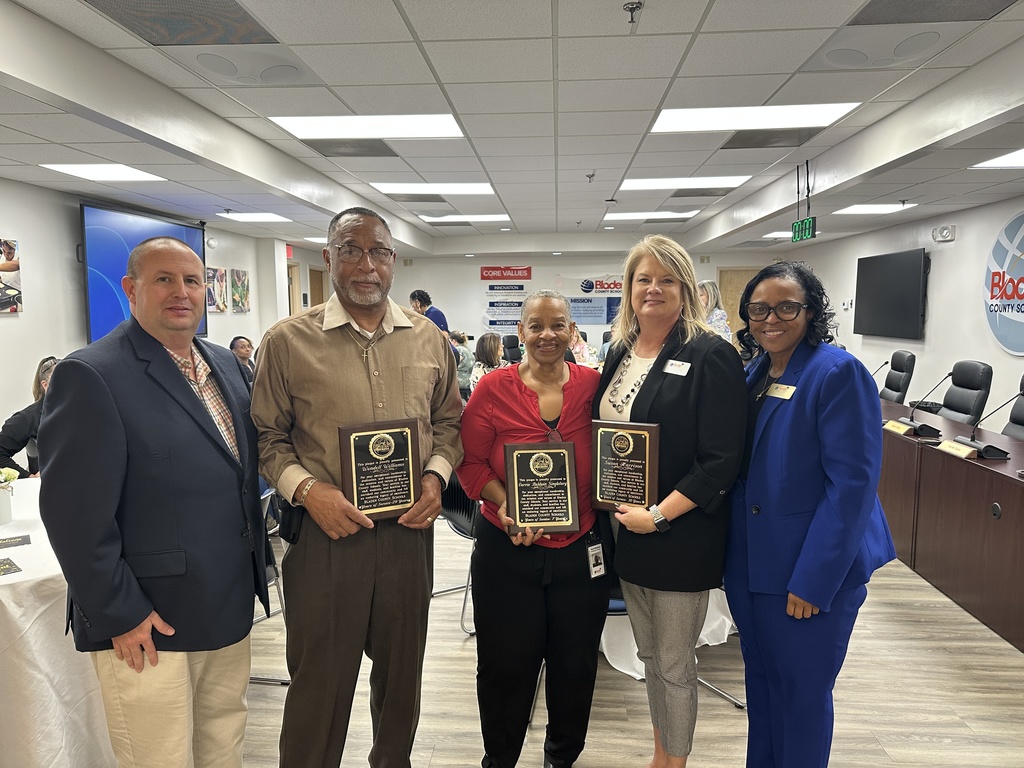 Group of five adults standing together in a meeting room, with three individuals in the center holding recognition plaques.