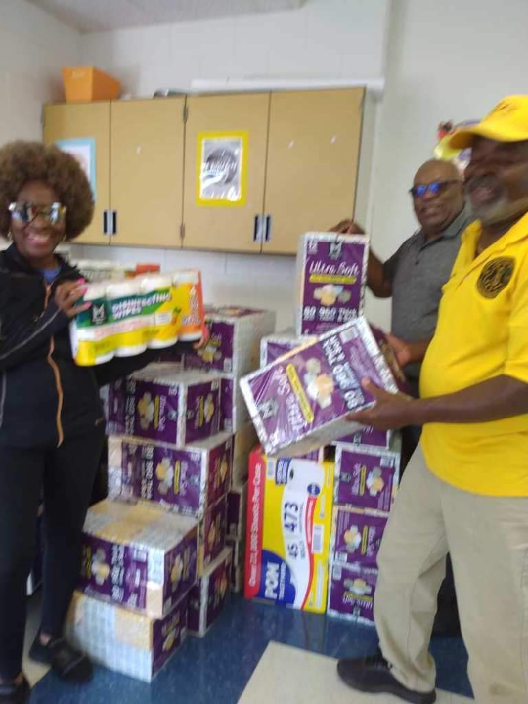 Three adults in a classroom unpack a large donation of paper towels and tissue boxes, stacking the supplies against a cabinet.