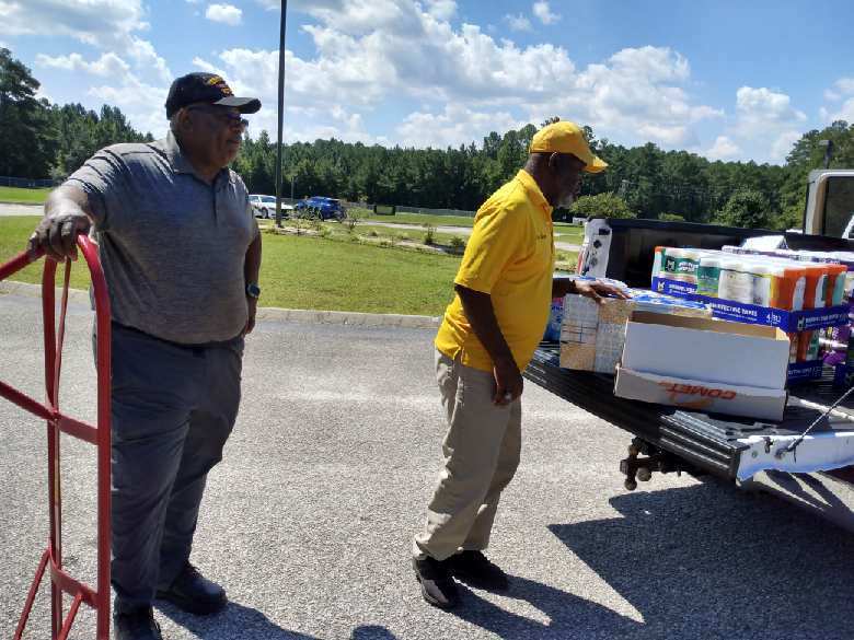 Two men stand beside a white pickup truck as they unload large boxes of paper towels and tissues in a school parking lot.