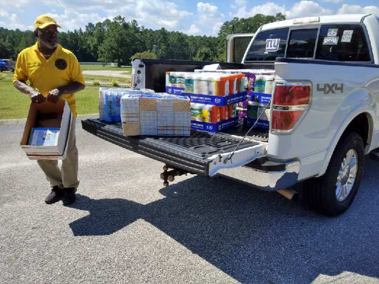 A man unloads boxes of paper towels and tissues from the bed of a white pickup truck parked outside a school.