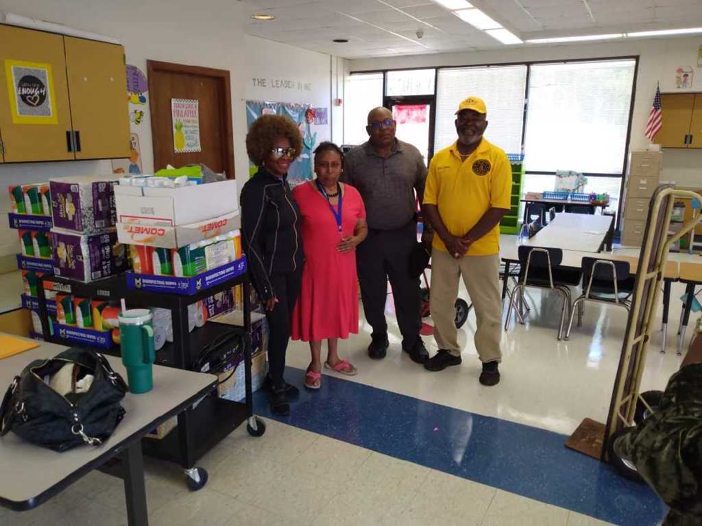 Four adults stand together in a classroom beside carts and stacks of donated paper towels, tissues, and cleaning wipes.