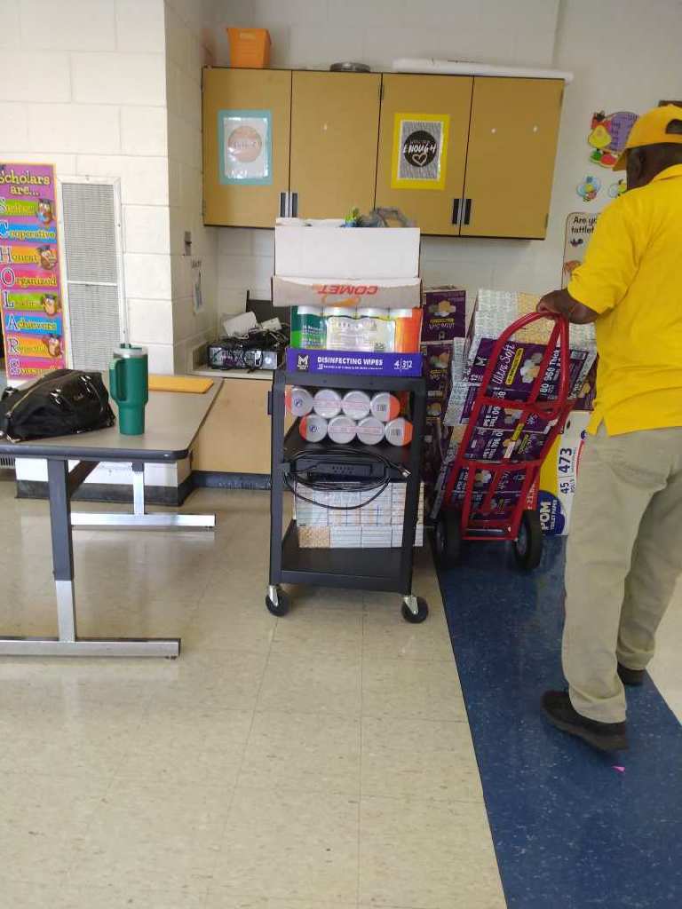 A man moves a red hand truck loaded with tissue boxes inside a classroom where donated supplies are stacked on a rolling cart.